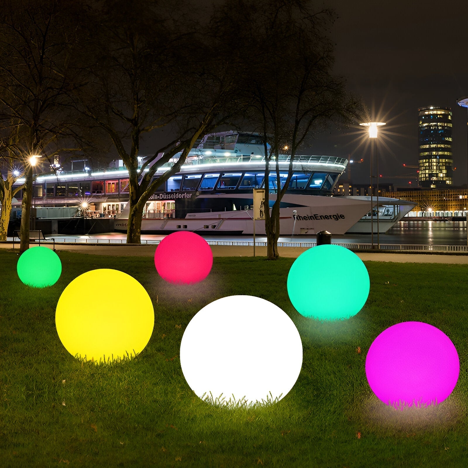 Colorful LED outdoor decorative spheres on grass at night, with a riverboat and city lights in the background. Perfect for garden or patio ambiance.