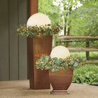 Outdoor LED decorative spheres with eucalyptus garland on tall bronze planters, perfect for garden lighting and patio decor, enhancing ambiance.