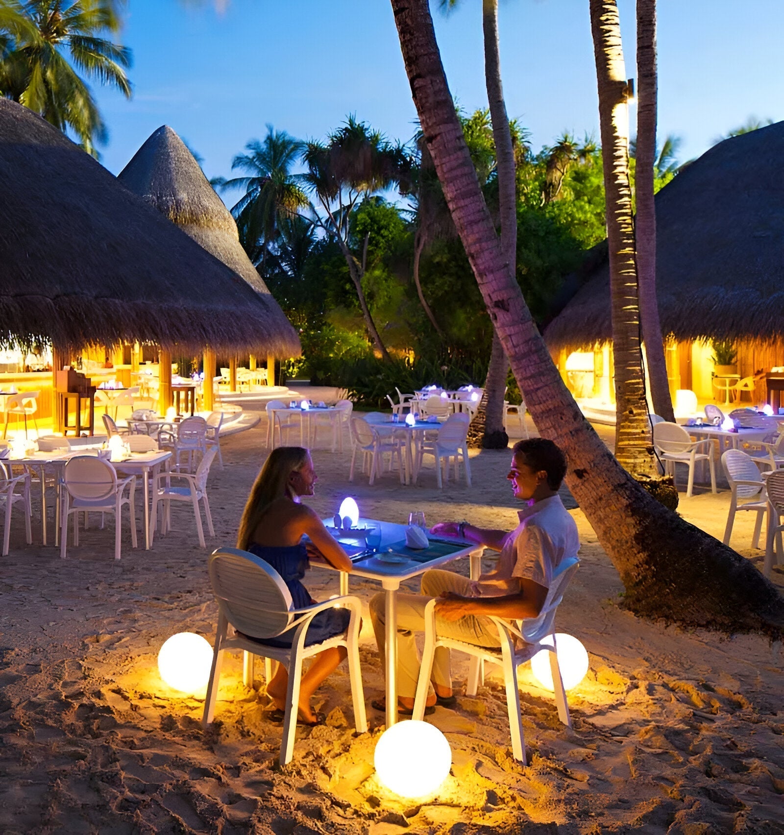 Couple dining on a tropical beach at sunset, surrounded by glowing LED sphere lights, palm trees, and thatched huts. Romantic outdoor ambiance.