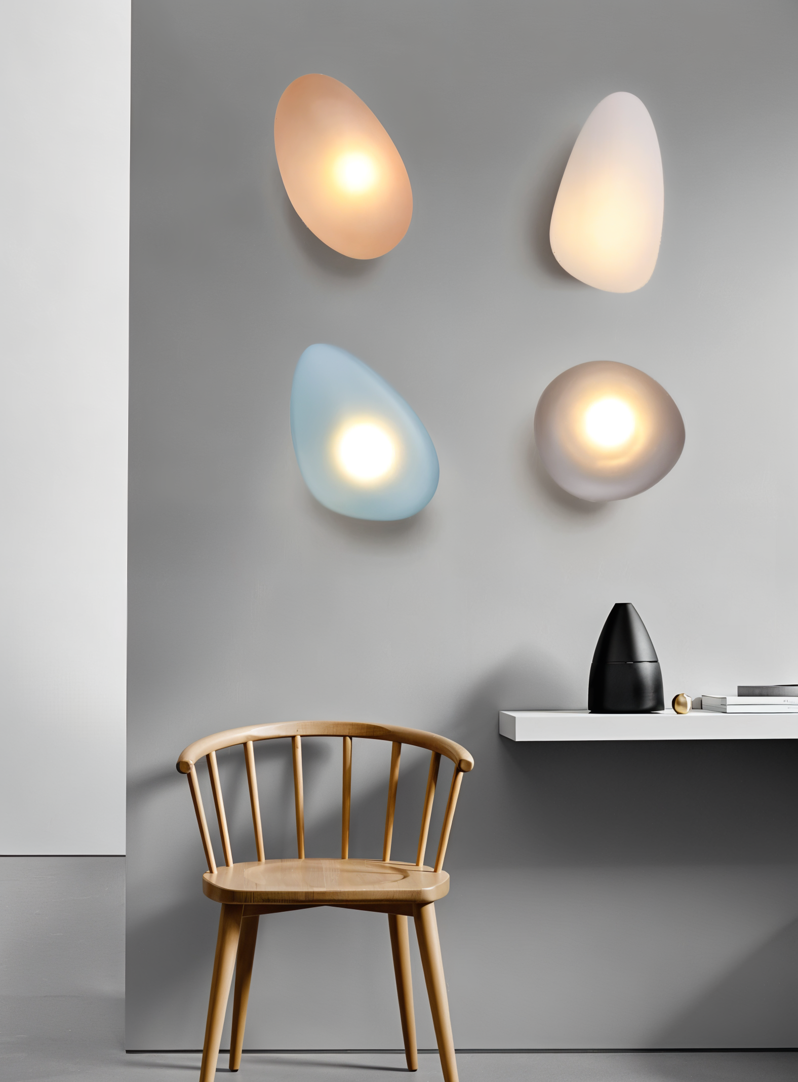 Modern pebble-shaped wall lights in soft hues on a gray wall, above a minimalist wooden chair and sleek white shelf. Contemporary home lighting decor.