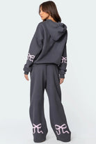 Woman wearing a dark gray hoodie and sweatpants set with pink bow designs on sleeves and legs, casual loungewear fashion, back view.