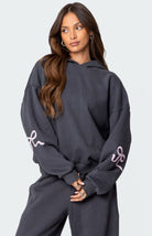 Woman wearing a dark gray hoodie with pink ribbon designs on sleeves, matching sweatpants, long brown hair, casual fashion, comfortable loungewear.