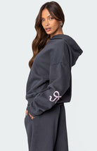 Woman wearing a dark gray hoodie with pink logo, matching sweatpants, and long brown hair. Fashionable casual wear, comfortable loungewear outfit.