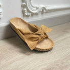 Tan suede bow knot sandal with cork sole on wooden floor, featuring elegant wall molding. Perfect for casual summer fashion and comfort.