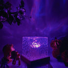 Illuminated glass block lamp with intricate patterns on a wooden base, casting vibrant purple and blue ambient light, surrounded by flowers and figurines.