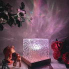 Illuminated glass block lamp with wooden base, surrounded by colorful light reflections, flowers, and toy figures. Modern home decor lighting.