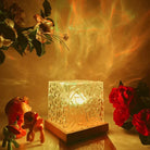 Illuminated glass block lamp with wooden base, surrounded by vibrant flowers and colorful figurines, creating a warm, artistic ambiance.