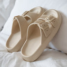 Beige slide sandals with double buckle straps on a white bed, featuring textured soles. Comfortable, stylish footwear for casual wear.