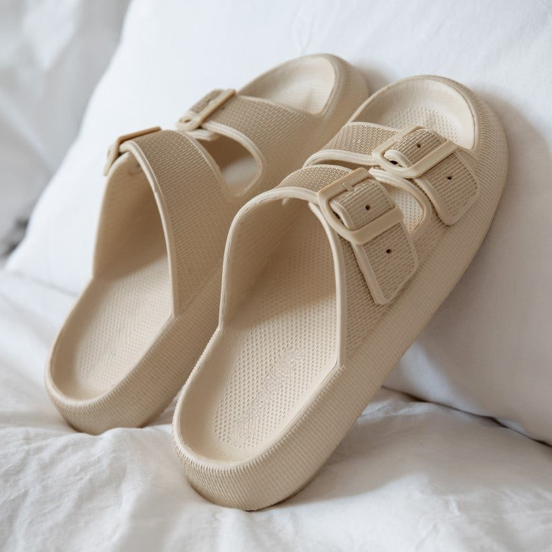 Beige slide sandals with double buckle straps on a white bed, featuring textured soles. Comfortable, stylish footwear for casual wear.