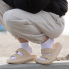 Beige slide sandals with double buckle straps worn with white socks, casual outdoor fashion, comfortable footwear, beach background.