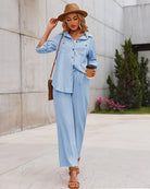 Woman in light blue button-up shirt and pants, wearing a brown hat and sandals, holding a coffee cup, walking outdoors. Fashionable casual outfit.