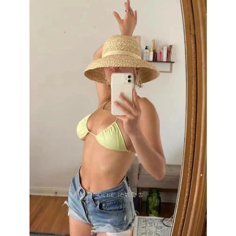 Woman in mirror wearing a wide-brimmed raffia hat, yellow bikini top, and denim shorts. Summer fashion, beachwear, straw hat, vacation style.