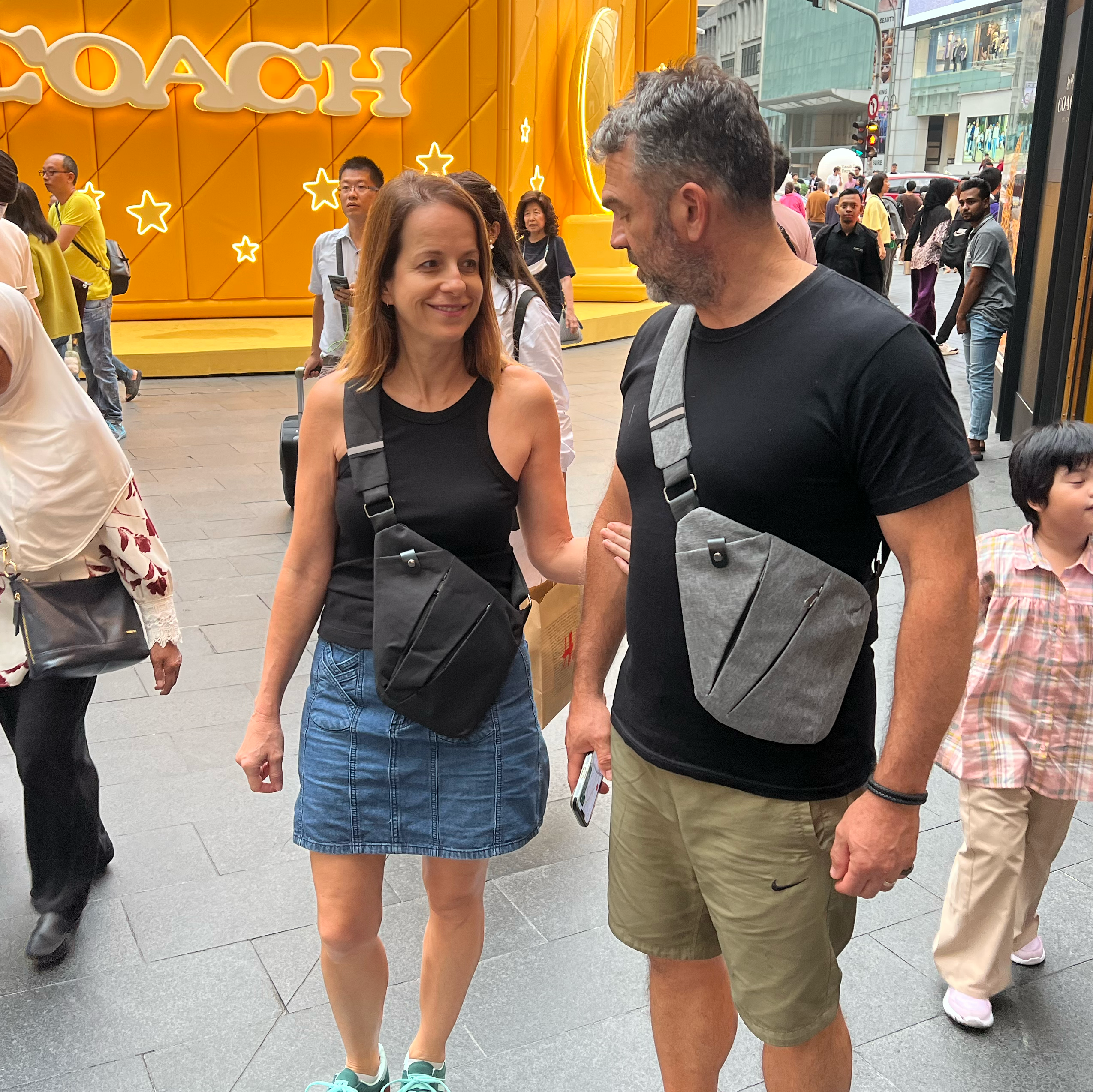 A couple wearing black t-shirts and crossbody bags walk in a busy shopping area. Bright yellow Coach store backdrop. Urban fashion, street style.