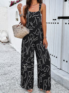 Woman wearing black abstract print jumpsuit with spaghetti straps, holding a woven straw handbag, standing against a white textured wall.