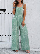 Woman wearing a green floral jumpsuit with white abstract patterns, standing against a white textured wall. Fashionable summer outfit, casual style.