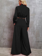 Woman in elegant black wide-leg jumpsuit with long sleeves, standing against a textured gray wall. Fashionable, modern women's clothing.