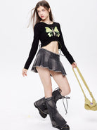 Young woman in a black butterfly crop top, gray pleated mini skirt, and oversized boots, holding a yellow textured handbag against a white background.