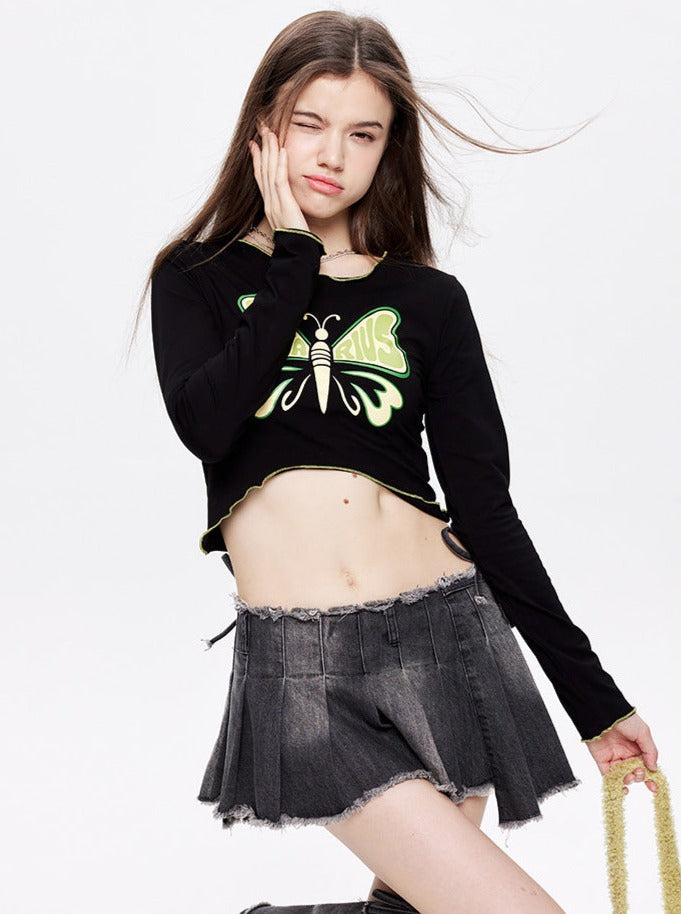 Young woman in a black long-sleeve crop top with a neon green butterfly design, paired with a frayed denim mini skirt, posing against a white background.