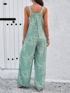 Woman wearing a green floral jumpsuit with white abstract patterns, standing against a white textured wall and door. Fashionable summer outfit.