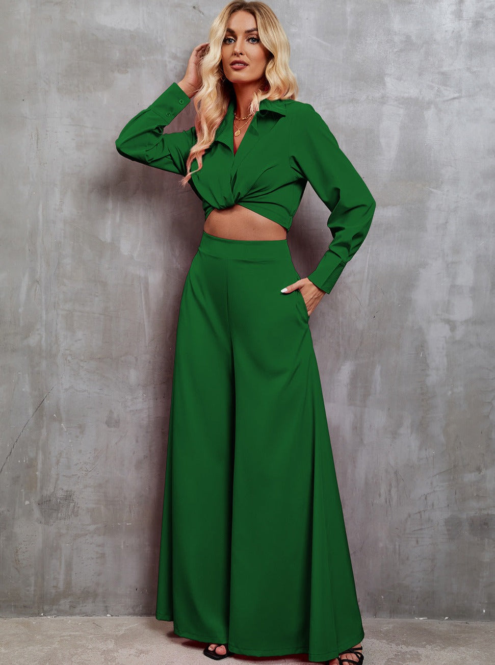 Woman in stylish green two-piece outfit with long sleeves and wide-leg pants, posing against a gray textured wall. Fashionable women's clothing.