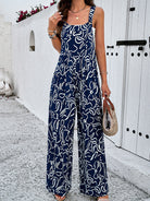 Woman in stylish blue and white abstract print jumpsuit, holding a woven handbag, standing against a white Mediterranean-style wall. Fashionable summer outfit.