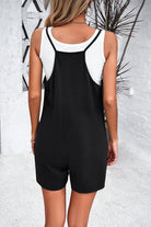 Woman wearing a black sleeveless romper over a white tank top, standing in a modern, minimalist setting. Fashionable summer outfit.