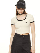 Woman wearing a black cap with text, cream crop top with black trim, and black pants. Fashionable casual outfit, modern streetwear style.