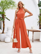 Woman in elegant orange halter-neck jumpsuit, holding a beige handbag, standing in a stylish outdoor setting with tropical plants and white decor.