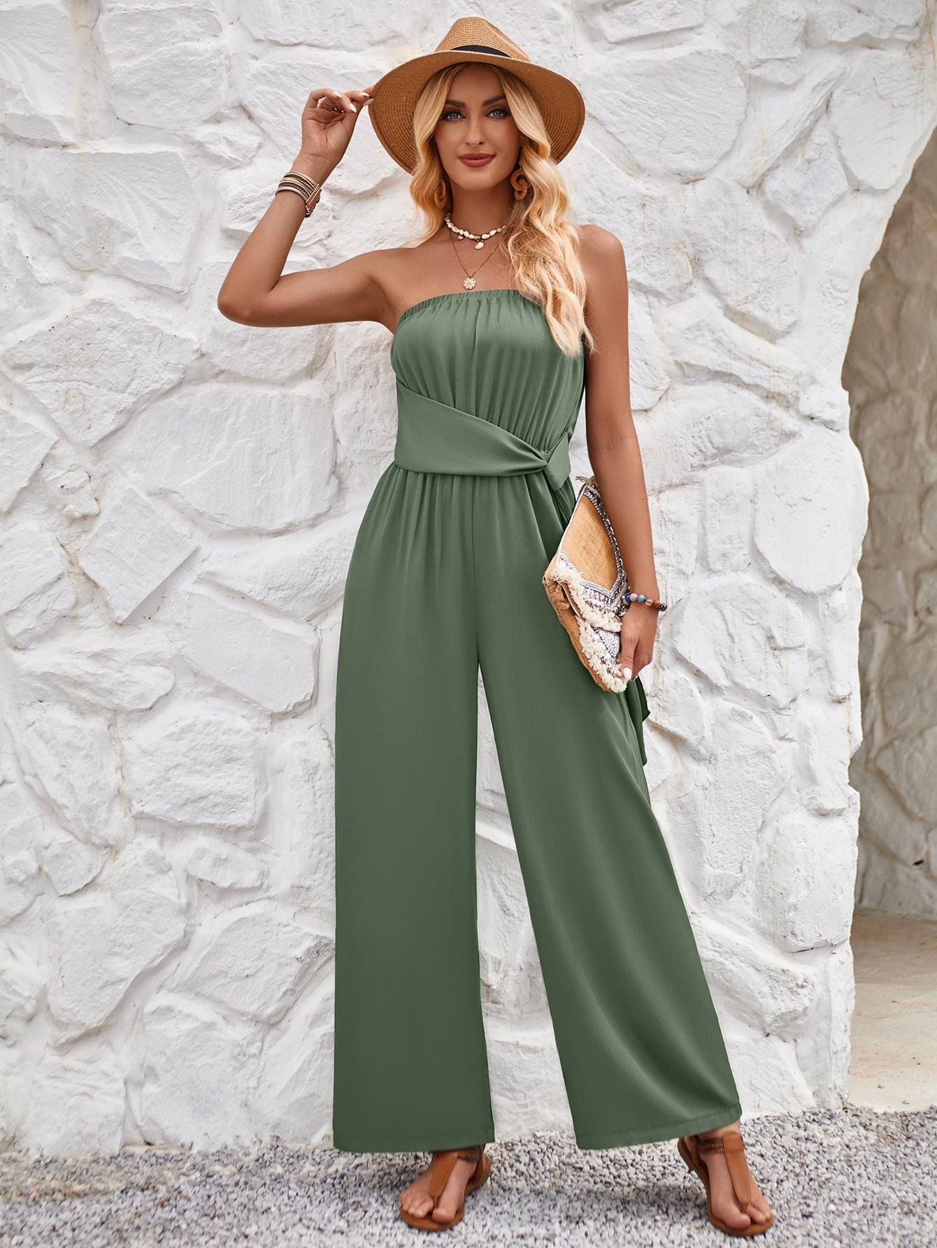 Woman in stylish green strapless jumpsuit, wearing a straw hat and holding a clutch, standing against a textured white stone wall. Fashionable summer outfit.