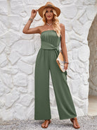 Woman in stylish green strapless jumpsuit, wearing a straw hat and holding a clutch, standing against a textured white stone wall. Fashionable summer outfit.