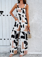 Woman wearing a stylish black and white geometric print jumpsuit, accessorized with bracelets and a straw handbag, standing against a textured wall.