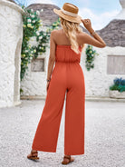 Woman in orange jumpsuit and straw hat, standing outdoors with white stone buildings and greenery. Summer fashion, casual style, vacation outfit.