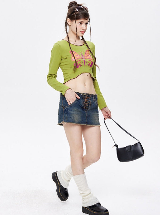 Young woman in a green crop top with butterfly design, denim mini skirt, black handbag, and boots. Fashionable Y2K style outfit, trendy streetwear.