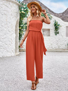 Woman in stylish orange jumpsuit and straw hat walking outdoors, summer fashion, casual chic outfit, white stone wall background, floral accents.