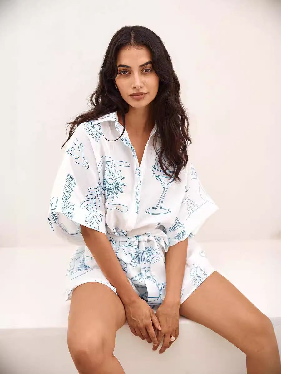 Woman wearing a stylish white romper with blue abstract patterns, sitting on a white bench. Fashionable summer outfit, casual chic style.