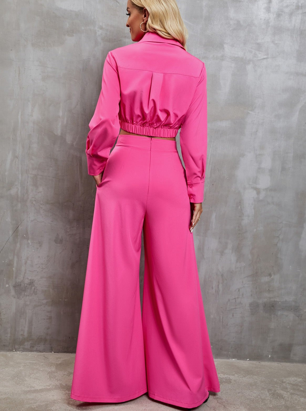 Woman in vibrant pink wide-leg jumpsuit with long sleeves, standing against a textured gray wall. Fashionable, stylish, modern women's clothing.