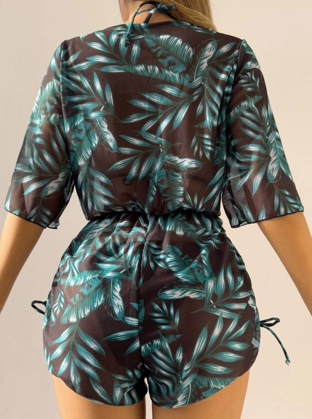 Woman wearing a tropical leaf print romper, featuring teal and black colors, short sleeves, and a cinched waist. Perfect for summer fashion trends.