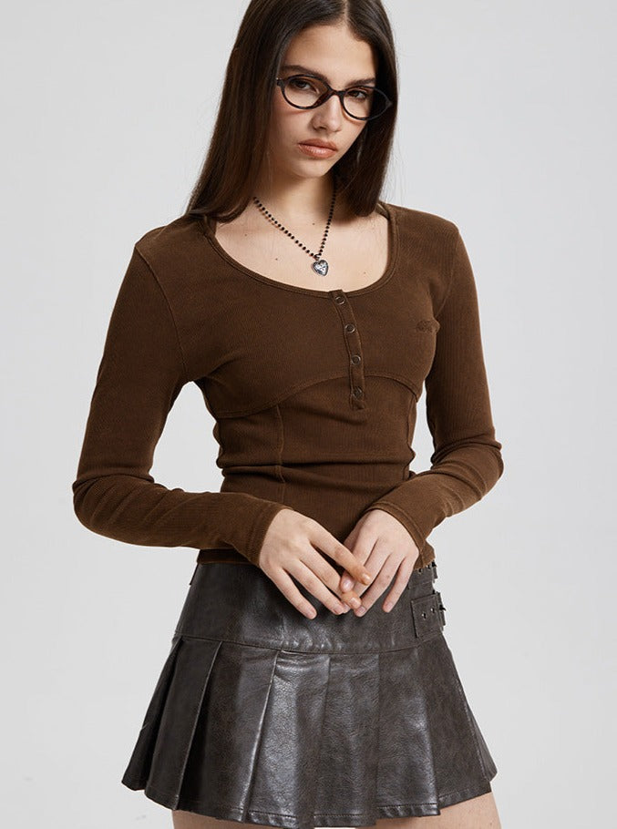 Woman in brown long-sleeve top and black leather pleated skirt, wearing glasses and a heart necklace, standing against a neutral background.