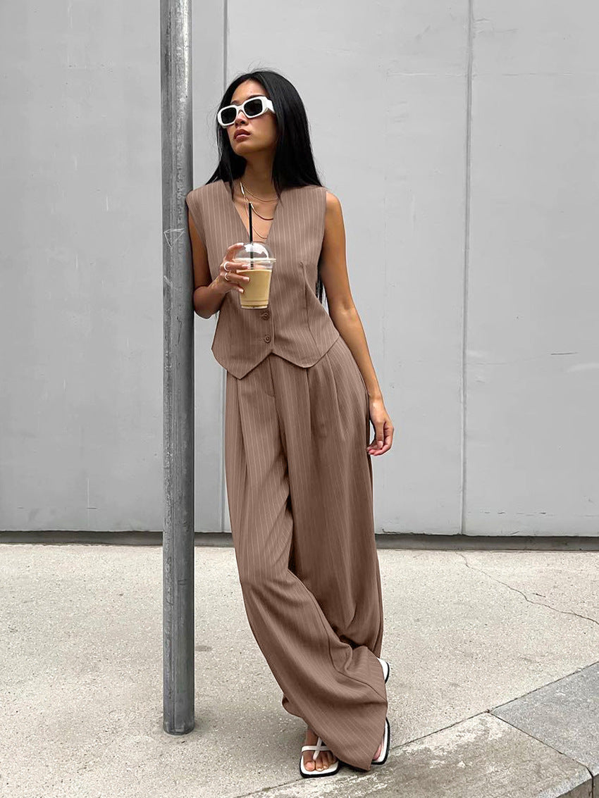 Woman in stylish brown pinstripe suit with wide-leg pants, holding iced coffee. Fashionable streetwear, sunglasses, urban setting.