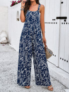 Woman in a stylish blue and white patterned jumpsuit, holding a woven handbag, standing on a white stone path. Fashionable summer outfit.