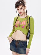 Young woman in a green long-sleeve crop top with a pink butterfly design, wearing a denim mini skirt and black shoulder bag, showcasing trendy fashion.