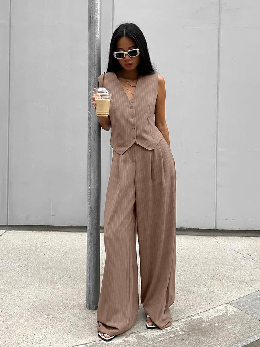 Woman in stylish brown pleated pantsuit, holding iced coffee, wearing white sunglasses and sandals, standing against a minimalist urban backdrop.