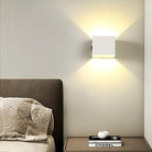 Modern wall sconce light fixture illuminating a minimalist bedroom with a gray upholstered headboard and a Chanel book on the nightstand.