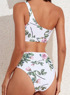 Woman wearing a floral one-shoulder bikini set with pink roses and green leaves, standing against a textured beige wall. Summer swimwear fashion.