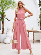 Woman in elegant pink halter-neck jumpsuit, holding a beige handbag, standing in a stylish outdoor setting with tropical plants and white decor.