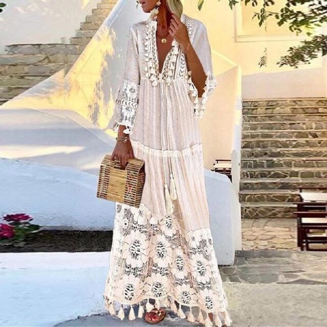 Woman in elegant white bohemian maxi dress with lace details, holding a wooden clutch, standing outdoors against a sunlit stone staircase backdrop.