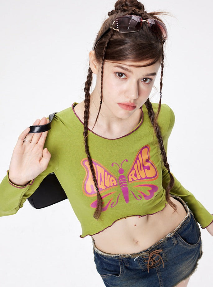 Young woman in green crop top with "Aquarius" butterfly design, wearing denim shorts and sunglasses on head, posing against white background. Fashion, trendy outfit.