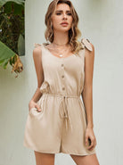 Woman wearing a stylish beige sleeveless romper with button front and tie waist, standing against a light background. Fashionable summer outfit.