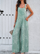 Woman wearing a stylish green floral jumpsuit with spaghetti straps, standing against a white textured wall. Fashionable summer outfit, casual chic.