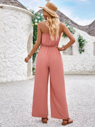 Woman in a pink sleeveless jumpsuit and straw hat, standing outdoors against a white stone wall. Fashionable summer outfit, casual style.